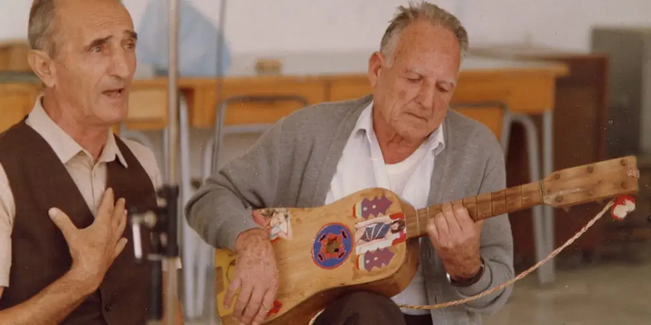 Due anziani suonano e cantano canzoni popolari italiane, uno con la mano sul cuore, l'altro con una chitarra decorata.