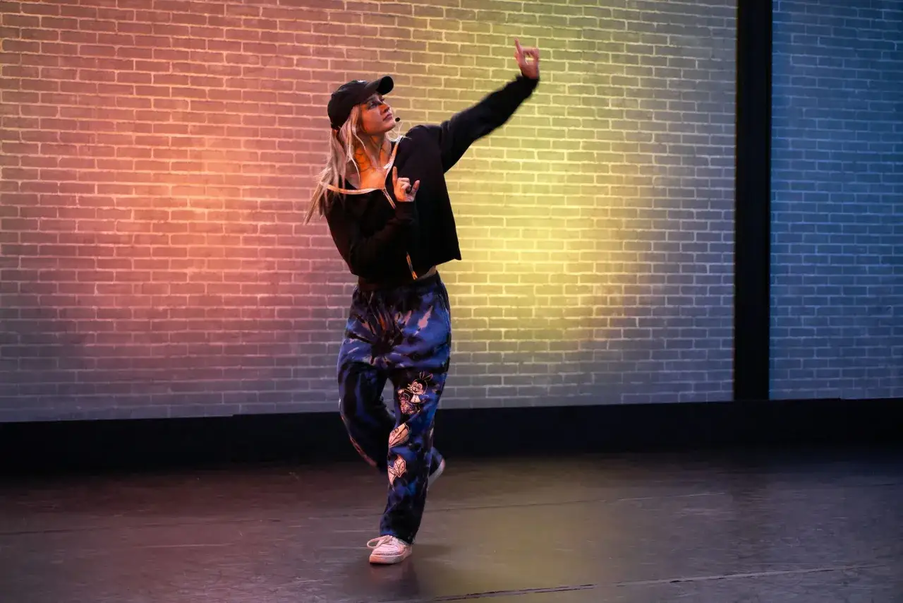 Ragazza con cappellino esegue una coreografia hip hop su un palco illuminato da luci colorate.