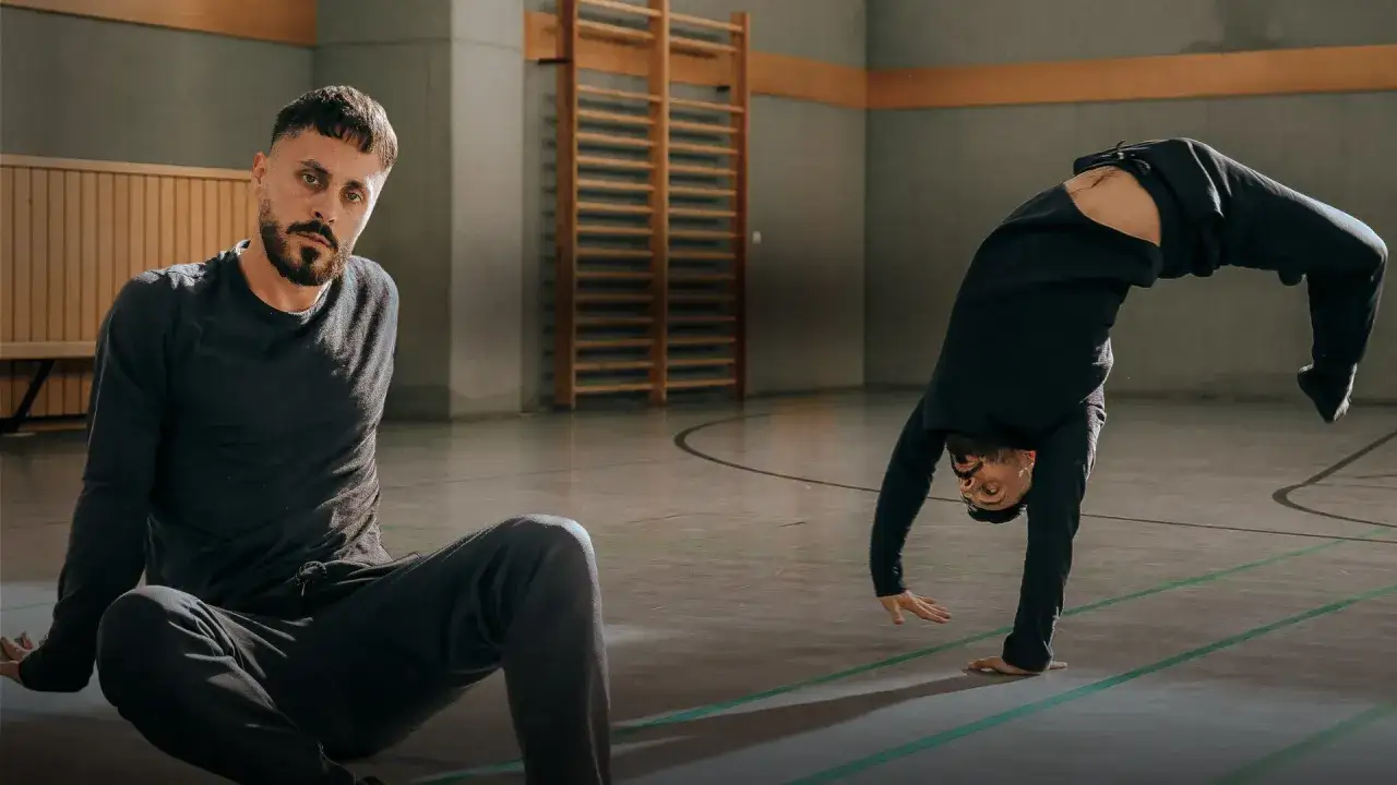 Due uomini in un ambiente sportivo, uno seduto con un'espressione intensa, l'altro in una posa acrobatica. Stile moderno contemporaneo.