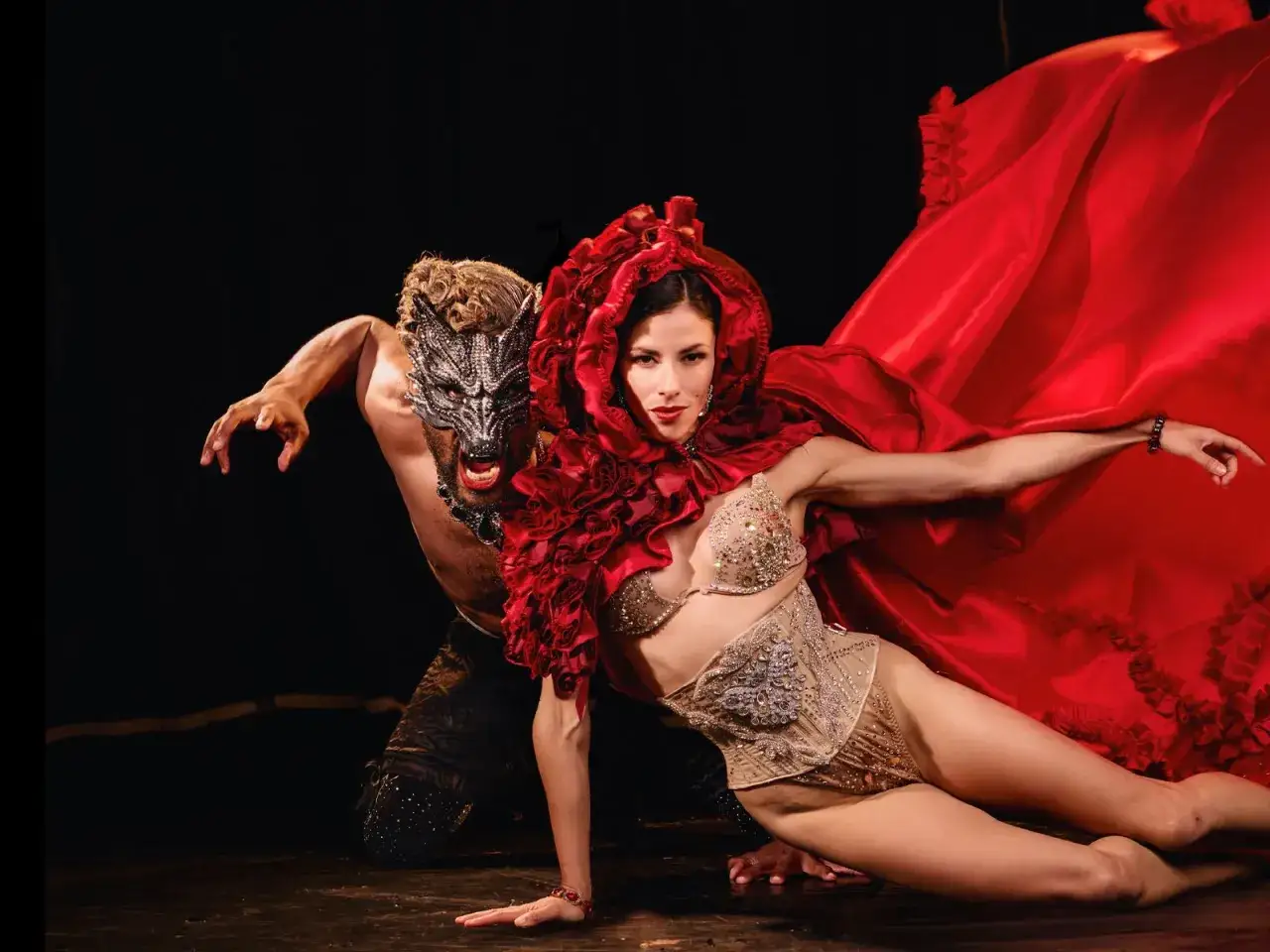 Coppia in un'esibizione di burlesque dance. Lei indossa un costume scintillante e un mantello rosso voluminoso, lui una maschera da lupo.