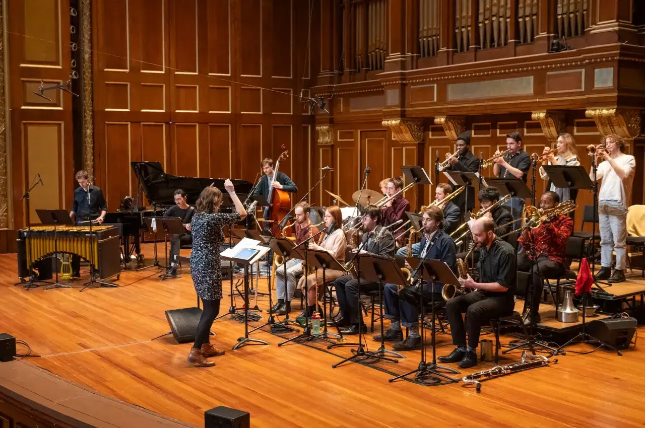 Un'orchestra di jazz suona sul palco, con fiati, ottoni e percussioni. La direttrice guida i musicisti.