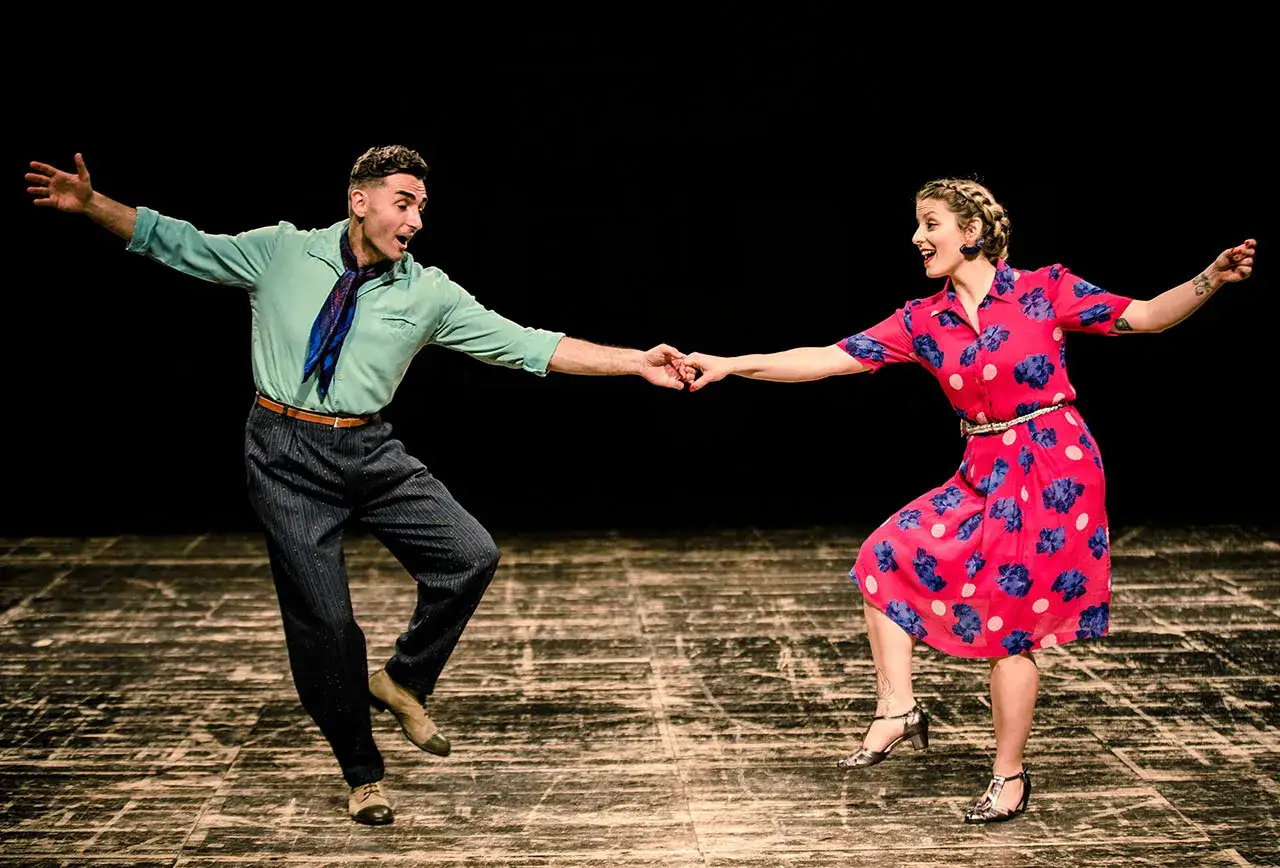 Coppia in abiti vintage esegue un energico lindy hop swing sul palco, con braccia aperte e sorrisi contagiosi.