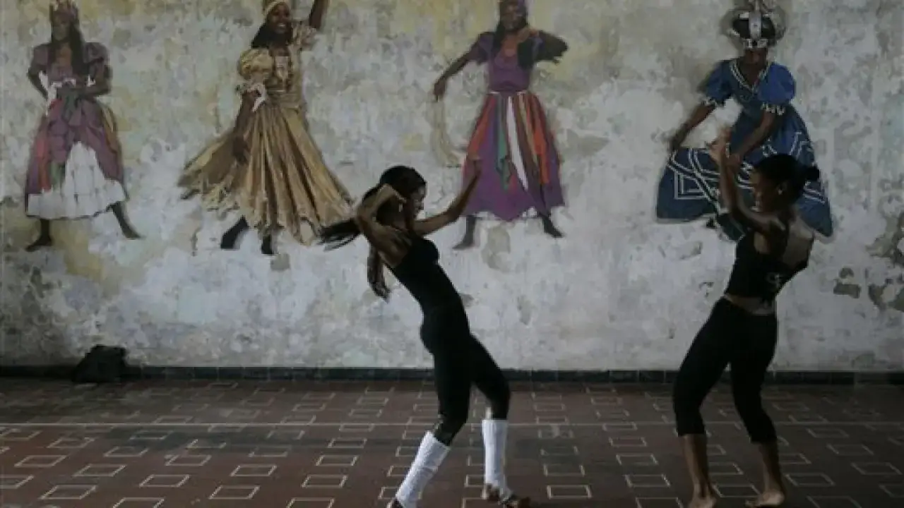 Due ballerine eseguono balli caraibici singoli davanti a un murale di figure femminili in abiti tradizionali.
