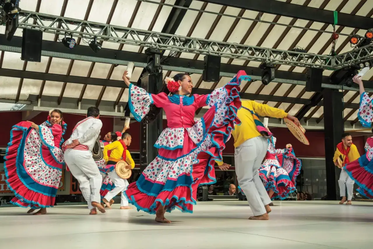 Danzatori in abiti colorati eseguono un vivace ballo americano, con gonne ampie e fiori tra i capelli.