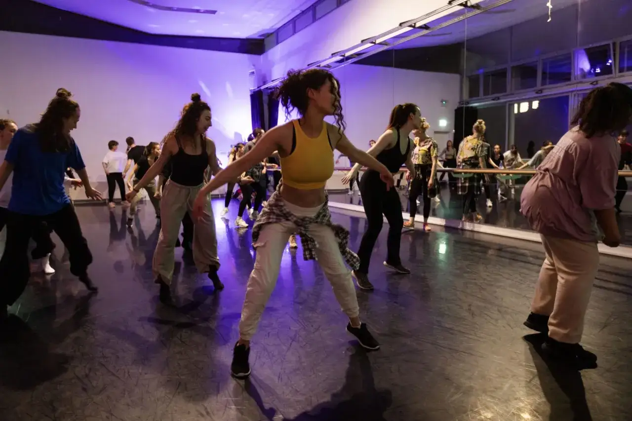 Un gruppo di ballerini si muove con energia in una sala prove illuminata da luci viola, praticando la danza reggaeton.