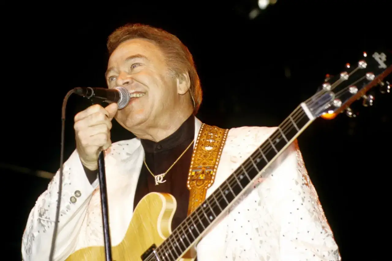 Un chitarrista famoso attuale, con giacca bianca ricamata, canta al microfono con la sua chitarra.