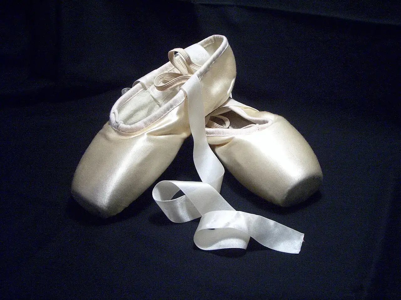 Un paio di punte da danza classica color crema con nastri bianchi drappeggiati su sfondo scuro.
