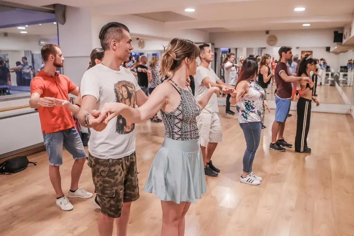 Coppie si esercitano in un corso di ballo latino americano, con mani intrecciate e passi coordinati su un pavimento di legno lucido.