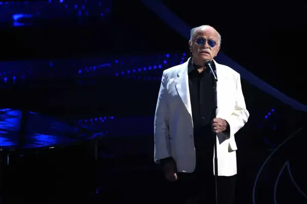 Un uomo con occhiali da sole blu canta sul palco, con un pianoforte a coda scuro e luci blu sullo sfondo. Sembra che sia lui chi ha scritto il cielo in una stanza.